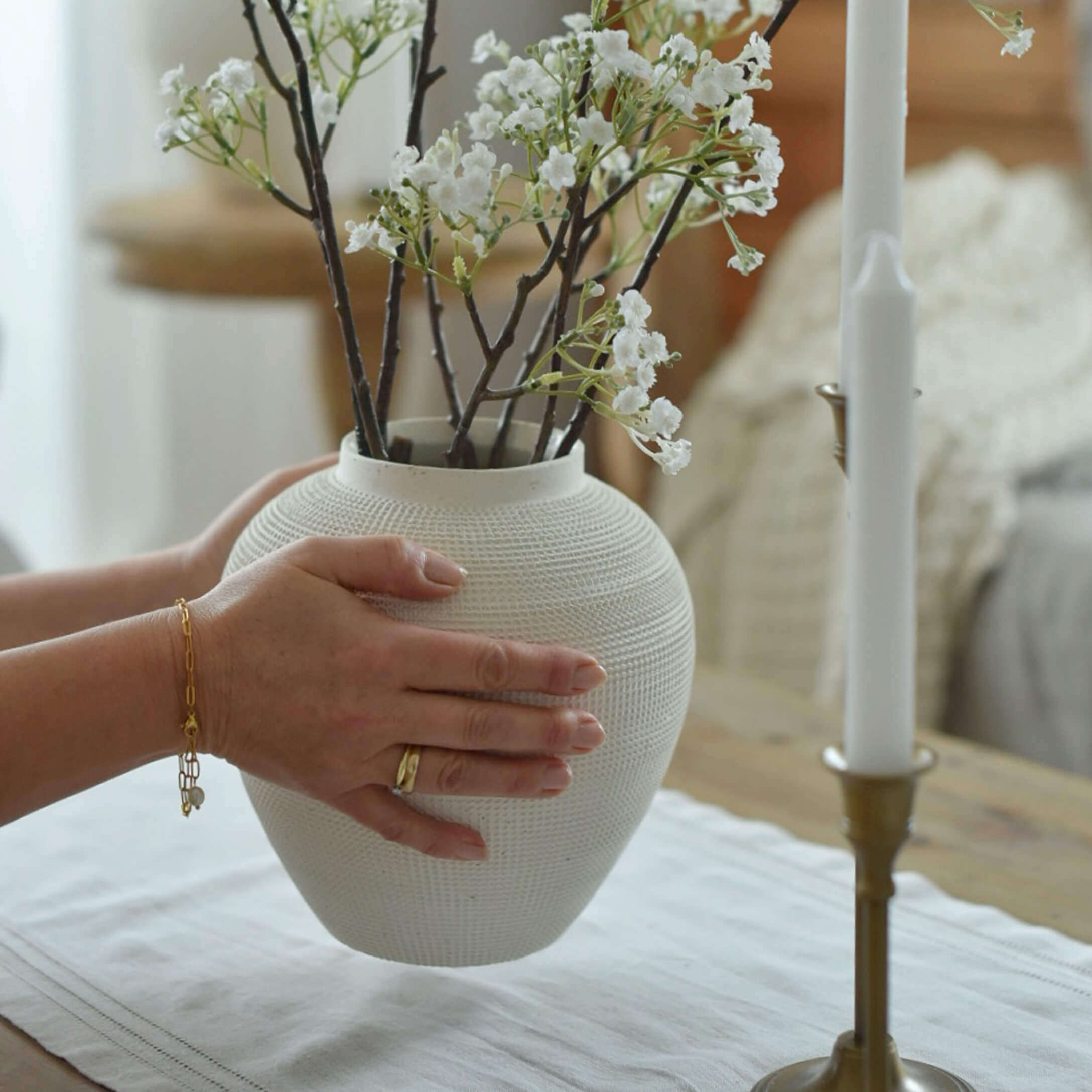 Vase en céramique blanche texturée tenu par des mains avec branches de petites fleurs blanches sur table