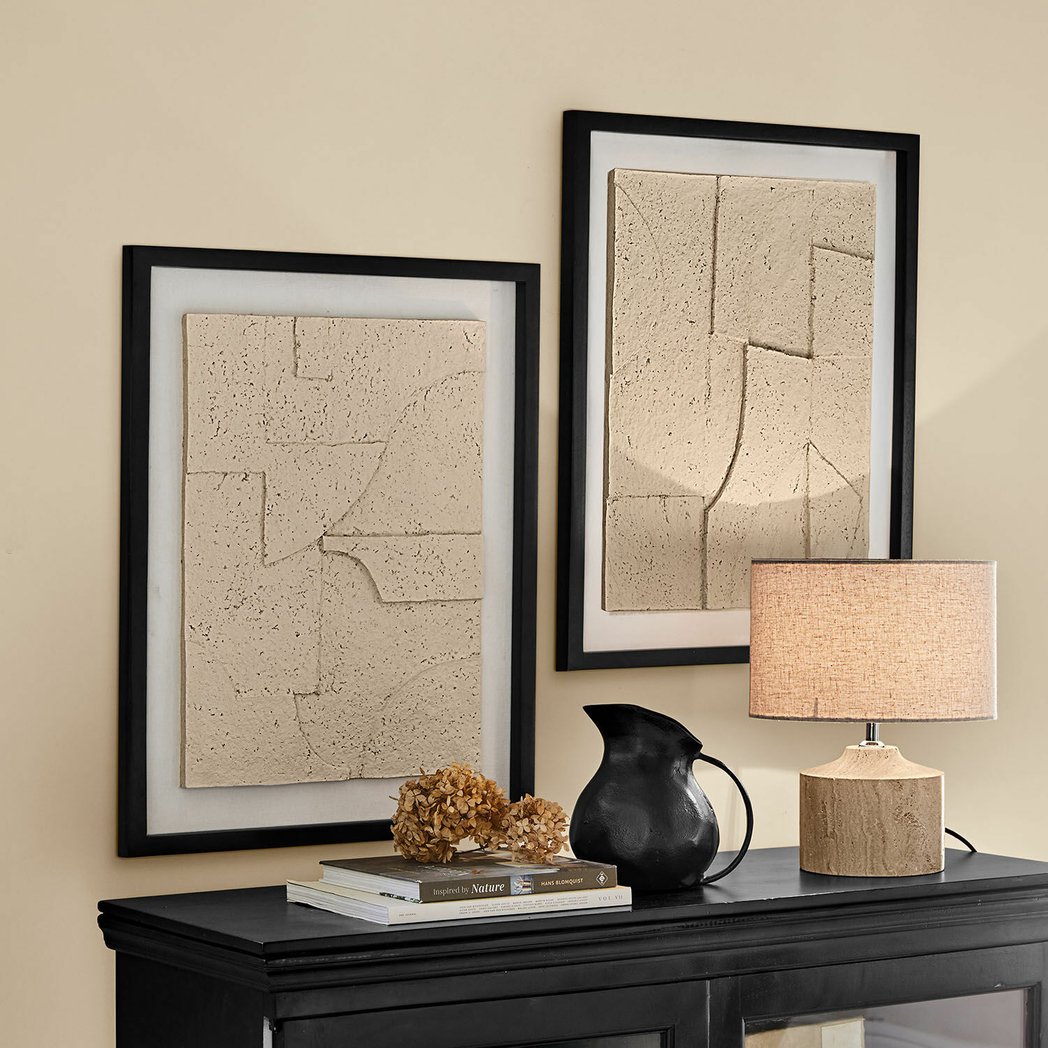 Lot de 2 tableaux Lavinois Deux tableaux en relief beige dans des cadres noirs au-dessus d'un buffet noir avec lampe et pichet noir