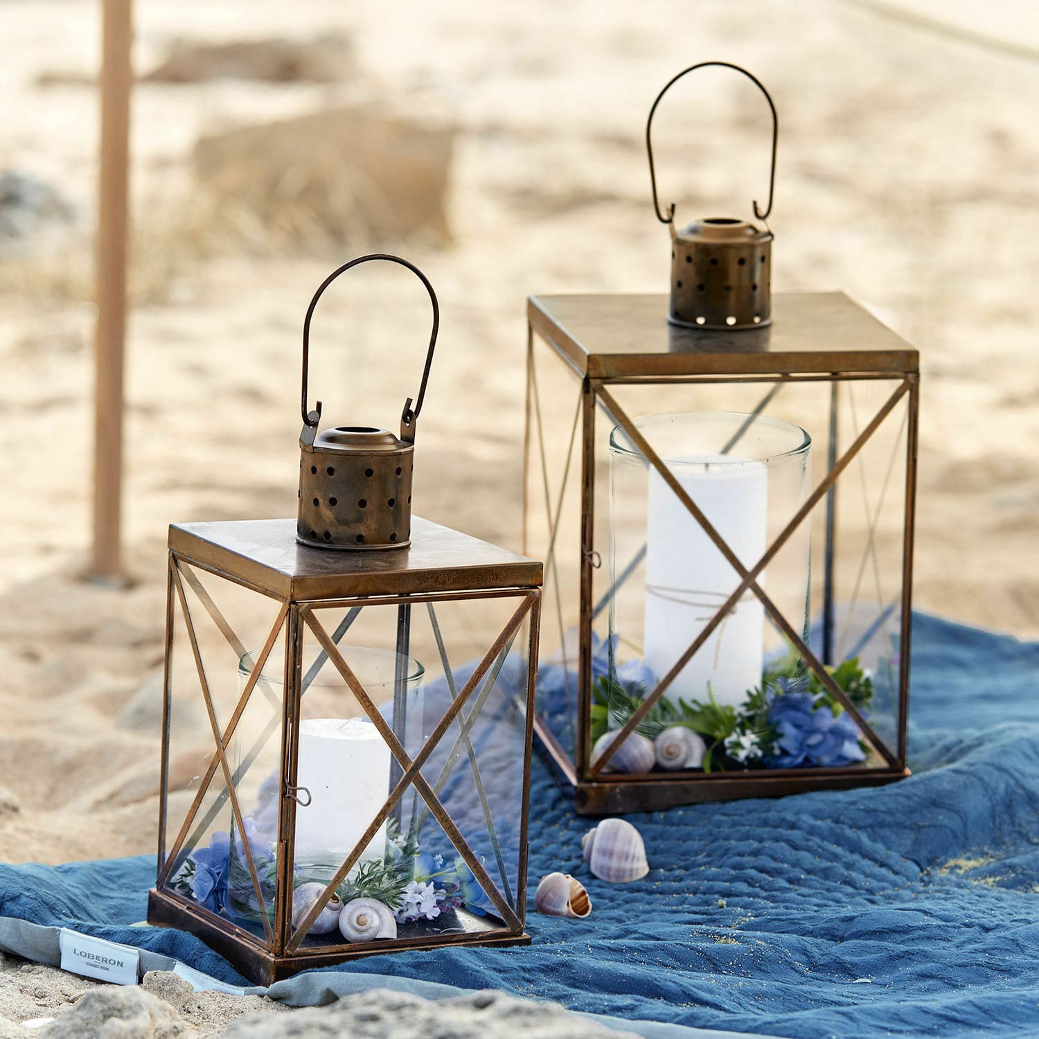 Lot de 2 lanternes Garance Deux lanternes en métal brun avec bougies blanches et décorations sur une plage sur une couverture bleue