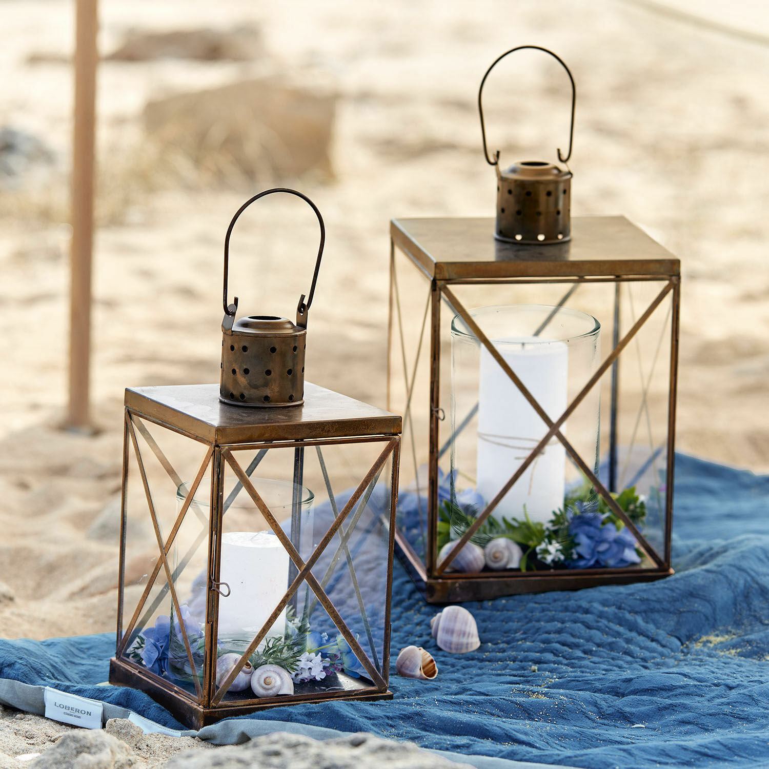 Lot de 2 lanternes Garance Deux lanternes en métal brun avec bougies blanches et décorations sur une plage sur une couverture bleue