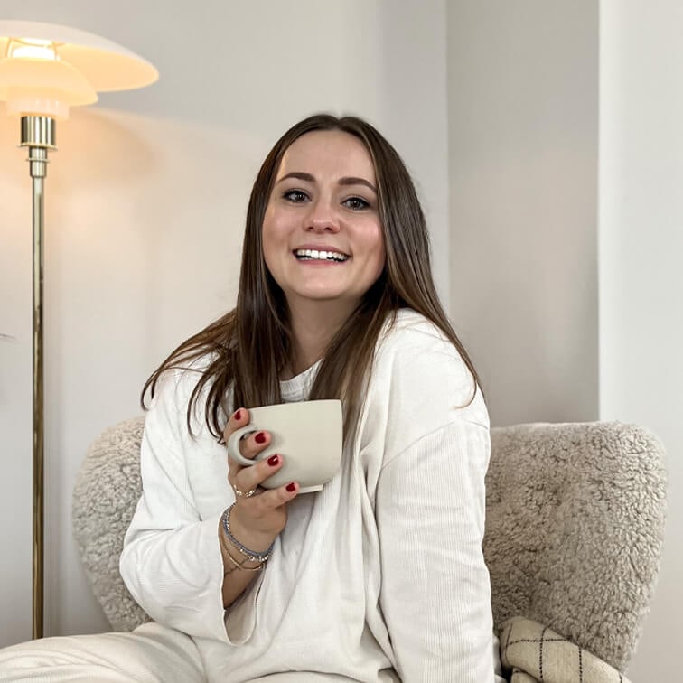 Femme souriante en pull blanc tenant une tasse beige assise sur un fauteuil moelleux dans un intérieur chaleureux