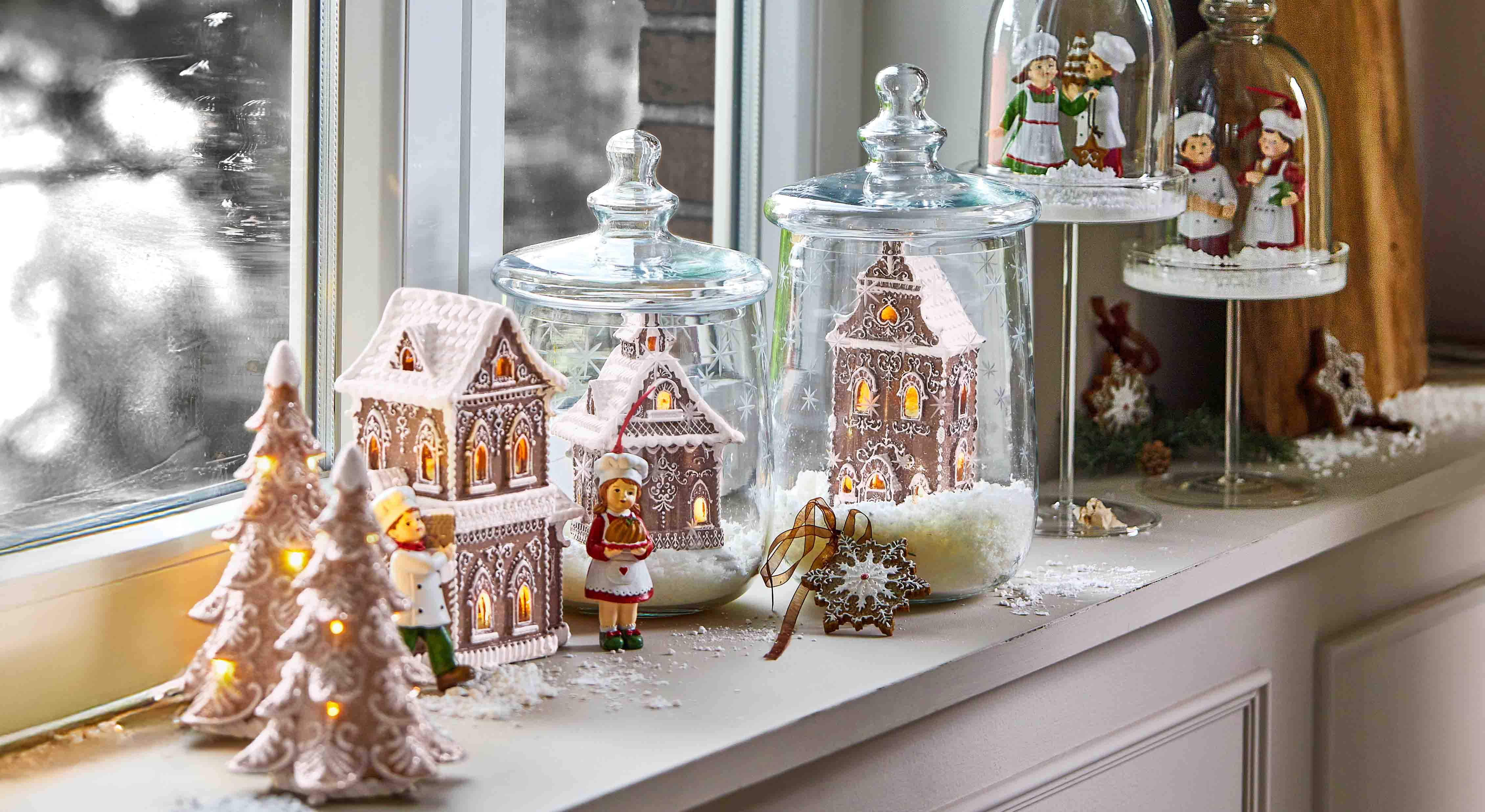 Maisons en pain d'épice décoratives et figurines de chefs sous cloches en verre sur un rebord de fenêtre avec éclairage hivernal