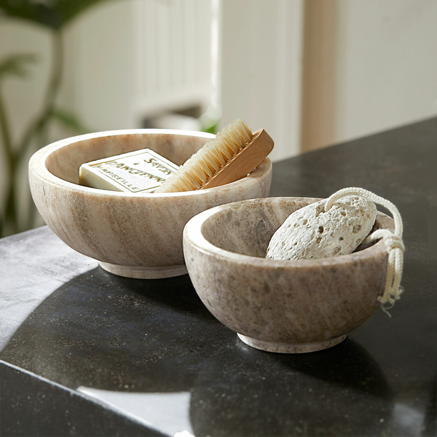 Lot de 2 coupelles Isamont Deux bols en marbre couleur naturelle avec savon, brosse et pierre ponce sur un comptoir noir de salle de bain
