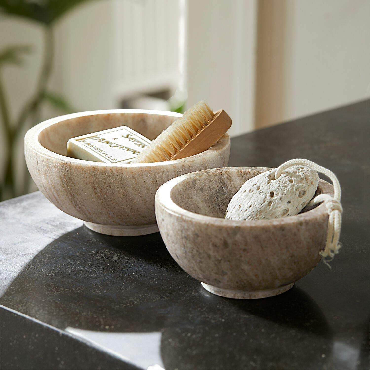 Lot de 2 coupelles Isamont Deux bols en marbre couleur naturelle avec savon, brosse et pierre ponce sur un comptoir noir de salle de bain