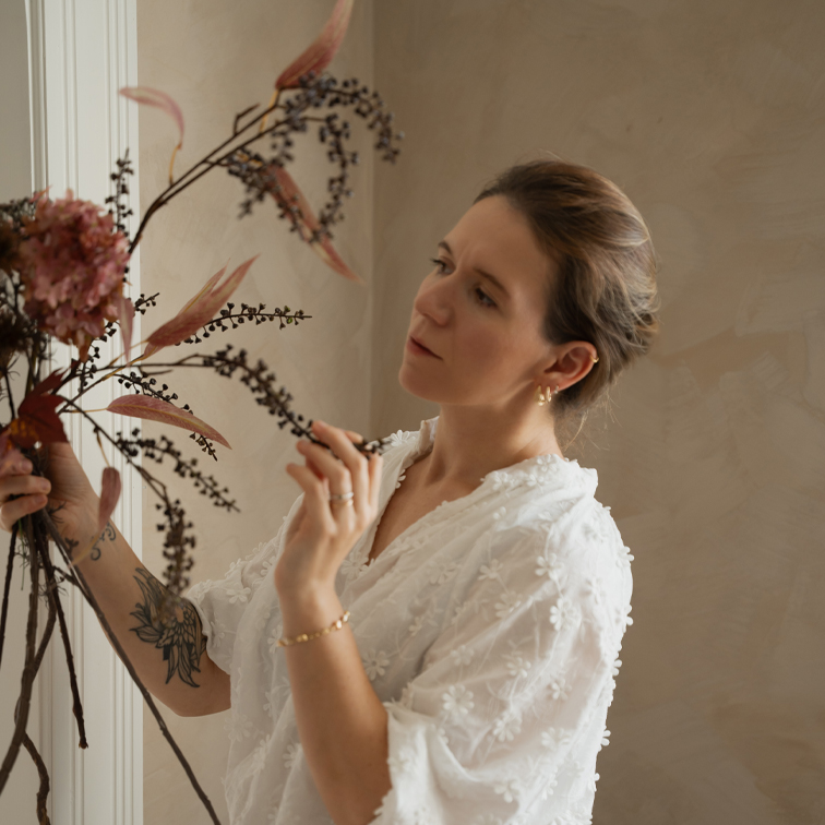 Femme en blouse blanche en dentelle arrangeant une décoration florale sèche dans un intérieur lumineux