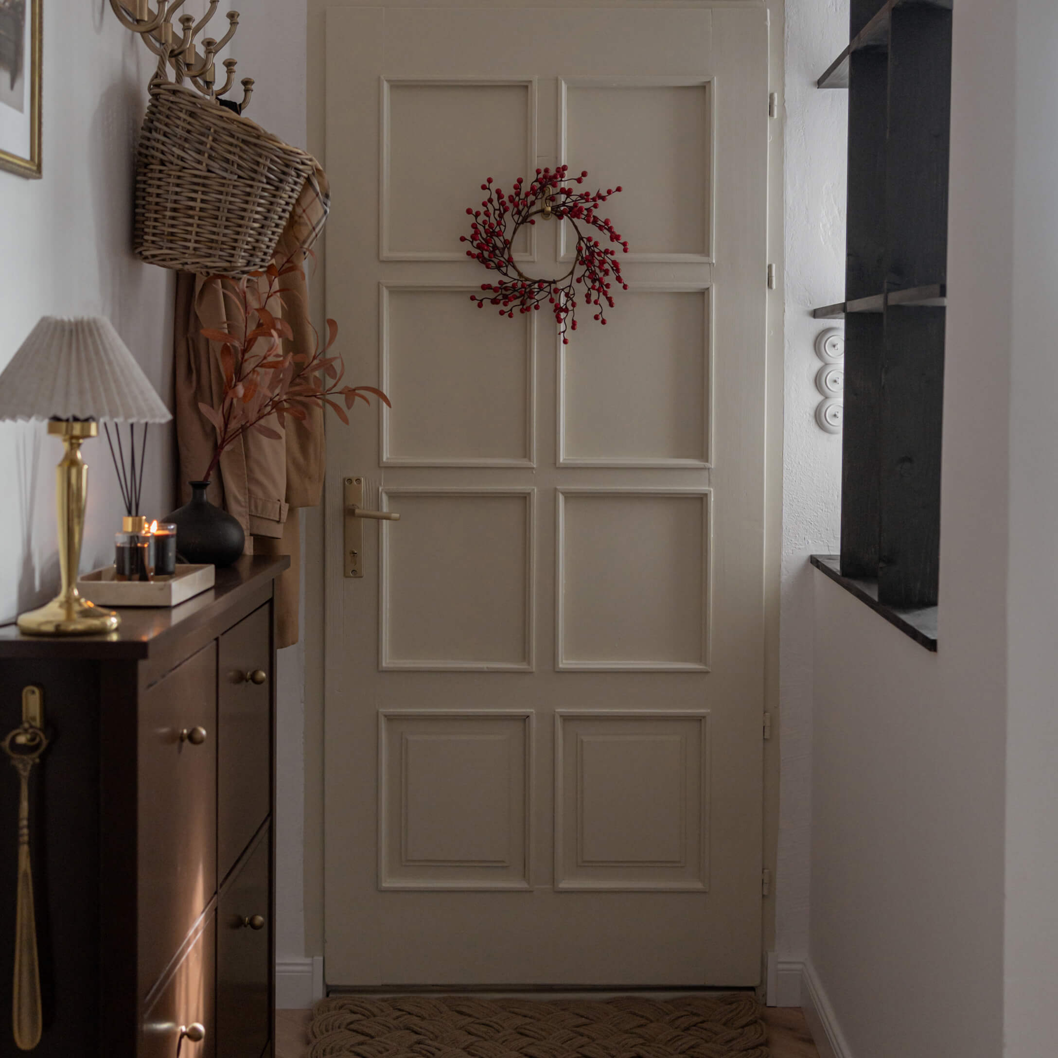 Entrée avec porte beige décorée d'une couronne rouge et meuble sombre avec lampe et bougies