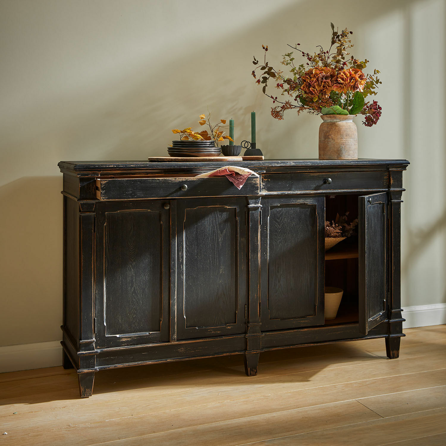 Buffet bas Chaveline Buffet en bois noir vieilli avec portes décoratives sur un sol en bois clair dans une pièce lumineuse.