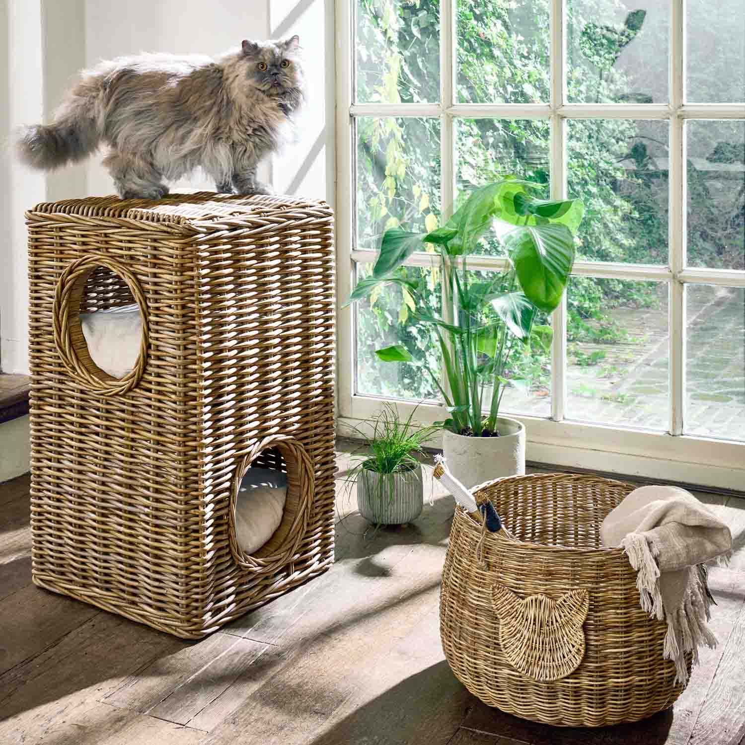 Maison pour chat en rotin naturel à deux niveaux avec coussins, panier et plantes près d'une fenêtre lumineuse