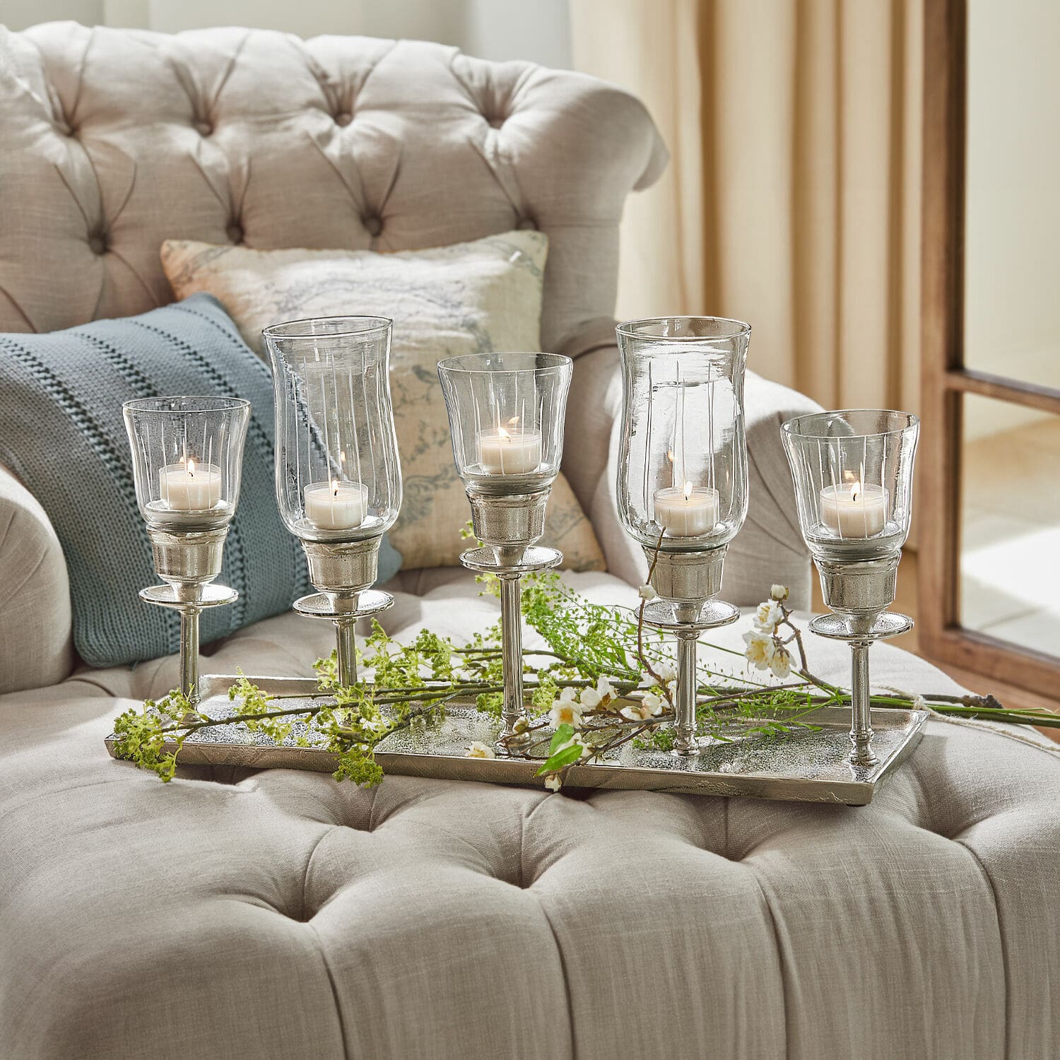 Plateau à bougies Ennedi Ensemble de cinq bougeoirs en verre et métal argenté posés sur un canapé beige avec coussins