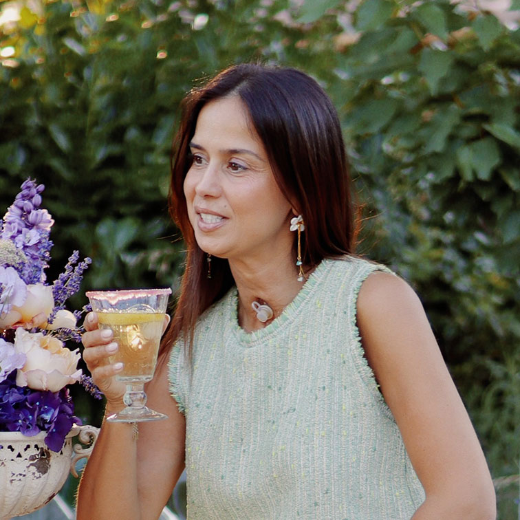 Femme en pull sans manches vert clair tenant un verre à boisson dans un jardin fleuri