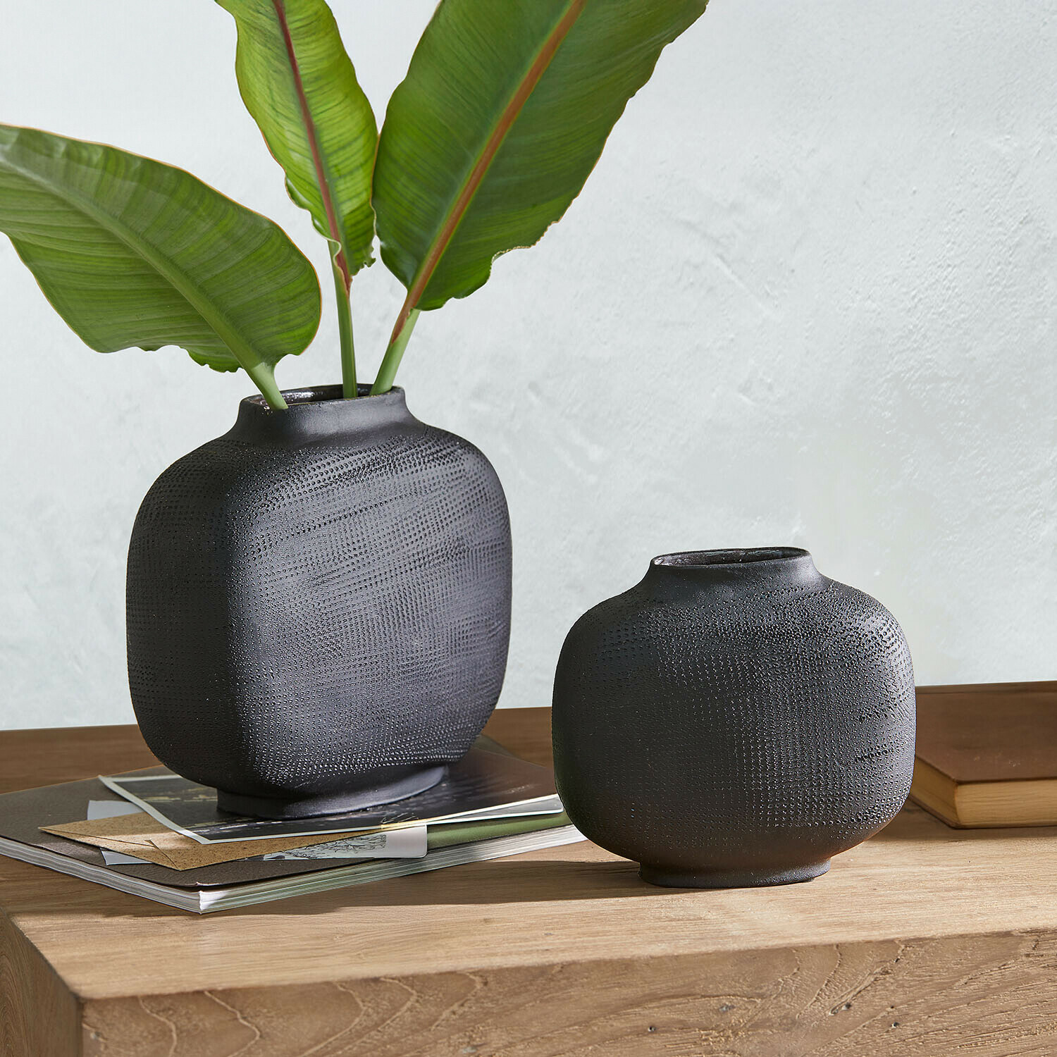 Deux vases en céramique noire mate avec texture posés sur une table en bois avec des feuilles vertes