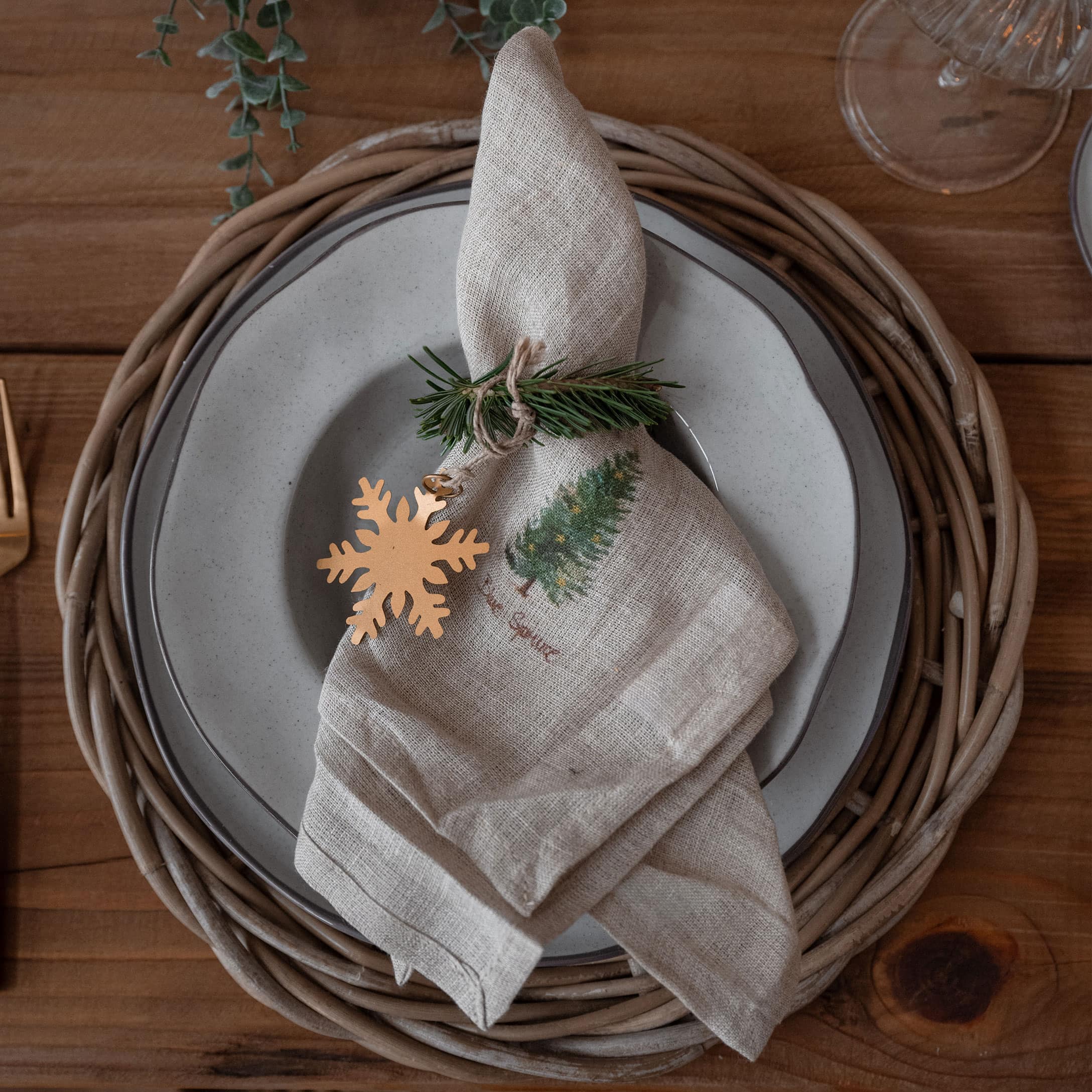 Serviette en lin avec motif sapin et décoration flocon sur assiette avec dessous en osier sur table en bois