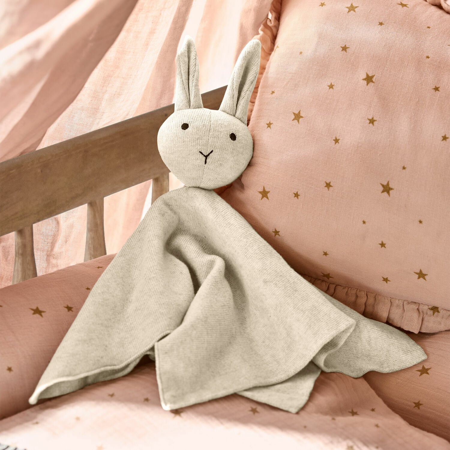 Doudou Lumiou Doudou lapin gris en tissu doux sur literie rose à étoiles, accessoire déco pour chambre d'enfant