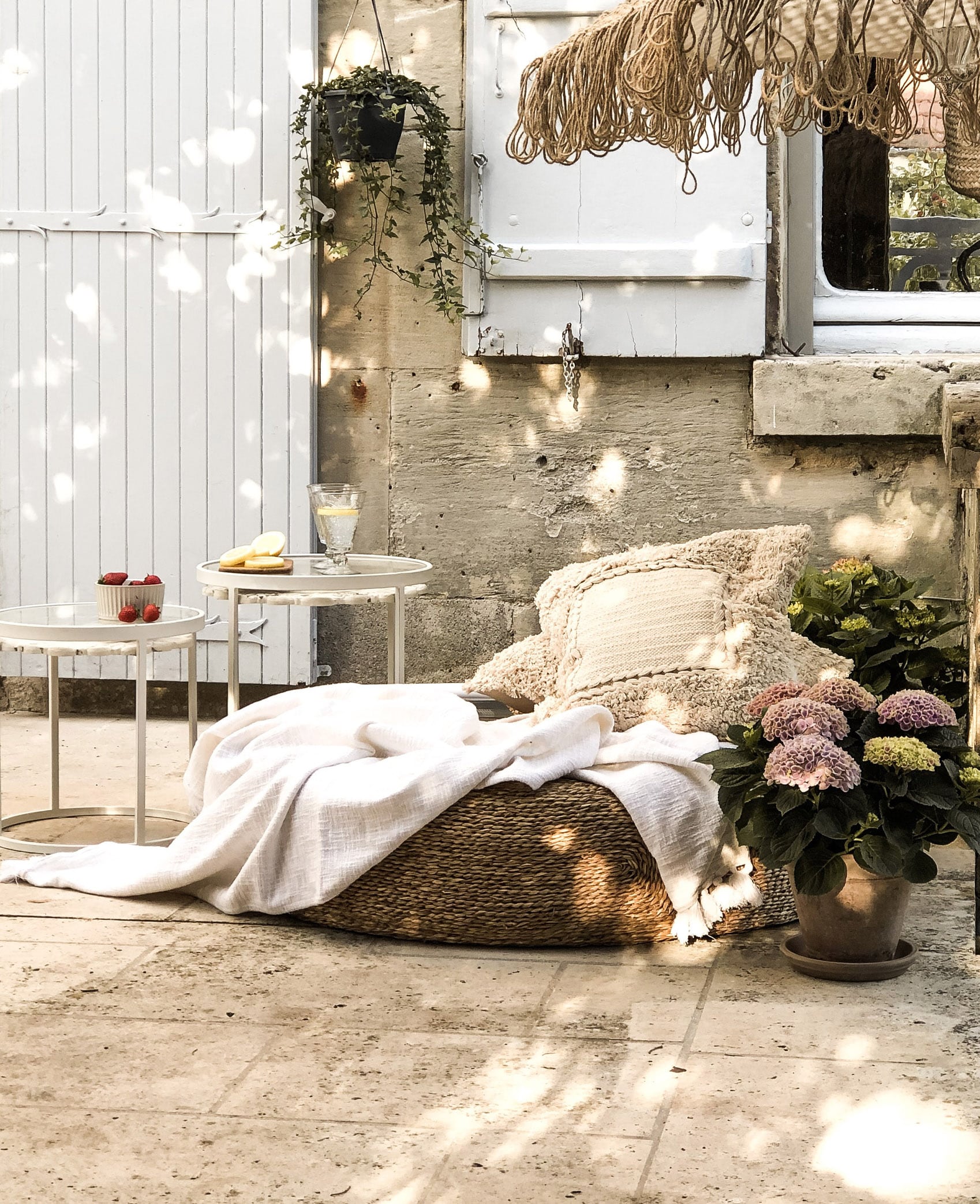 Coin cosy avec pouf en osier, couverture crème et coussin sur terrasse en pierre avec fleurs en pot.