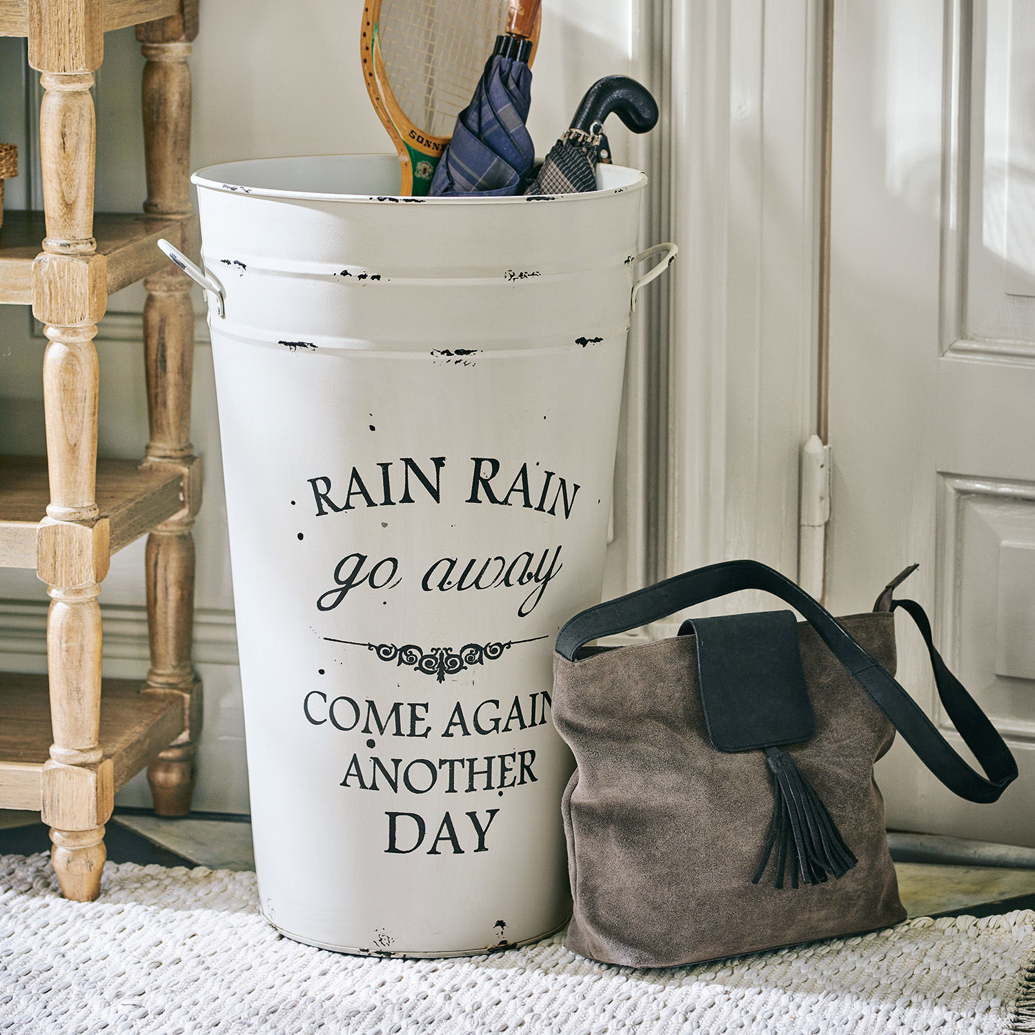Porte-parapluies Rain Porte-parapluie en métal blanc avec inscription et sac en daim marron posé au sol près de la porte