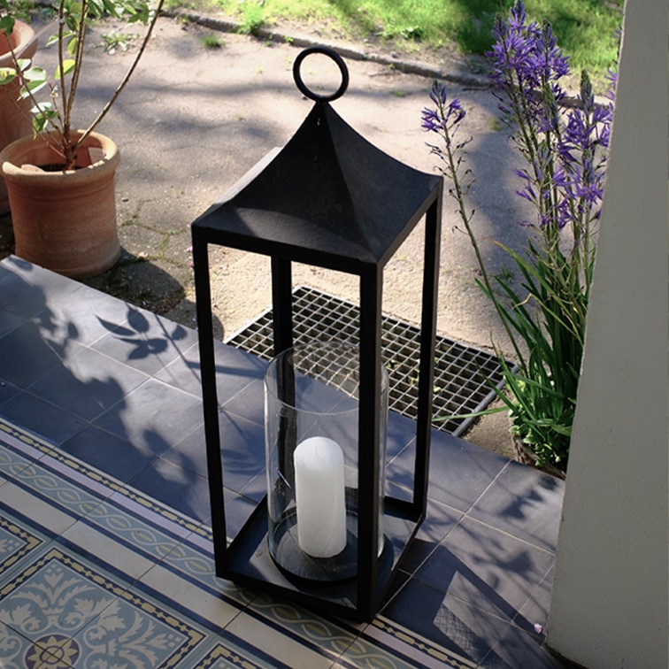 Lanterne noire en métal avec bougie blanche et verre protecteur sur terrasse carrelée avec plantes