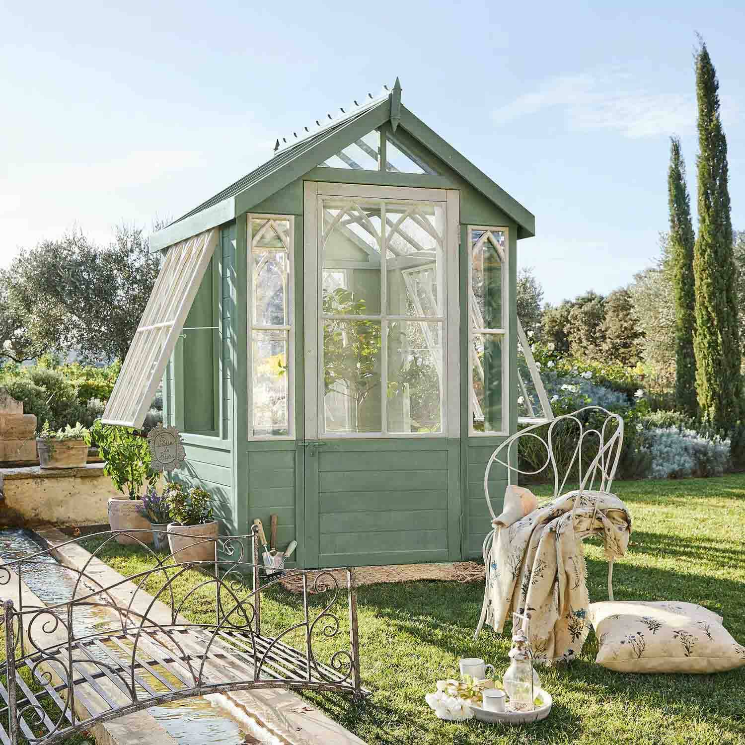 Serre en bois vert avec fenêtres blanches dans un jardin, chaise en métal et coussin à côté
