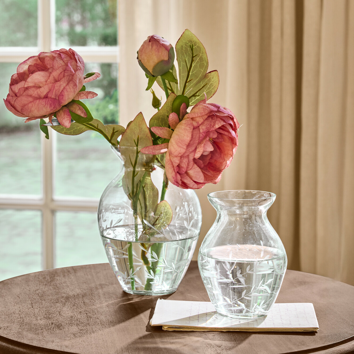 Vases en verre transparent avec motif gravé de feuilles, l'un avec des fleurs roses, sur une table en bois