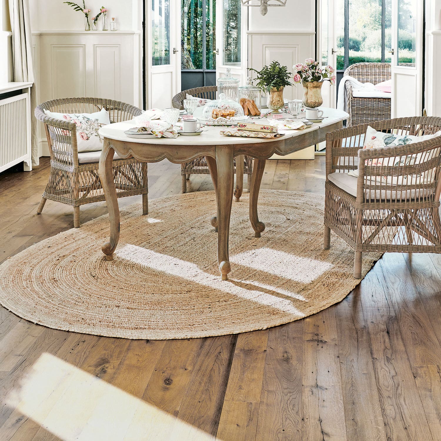 Tapis Ausone Table en bois clair avec plateau blanc sur tapis rond en jute dans un salon lumineux avec chaises en osier