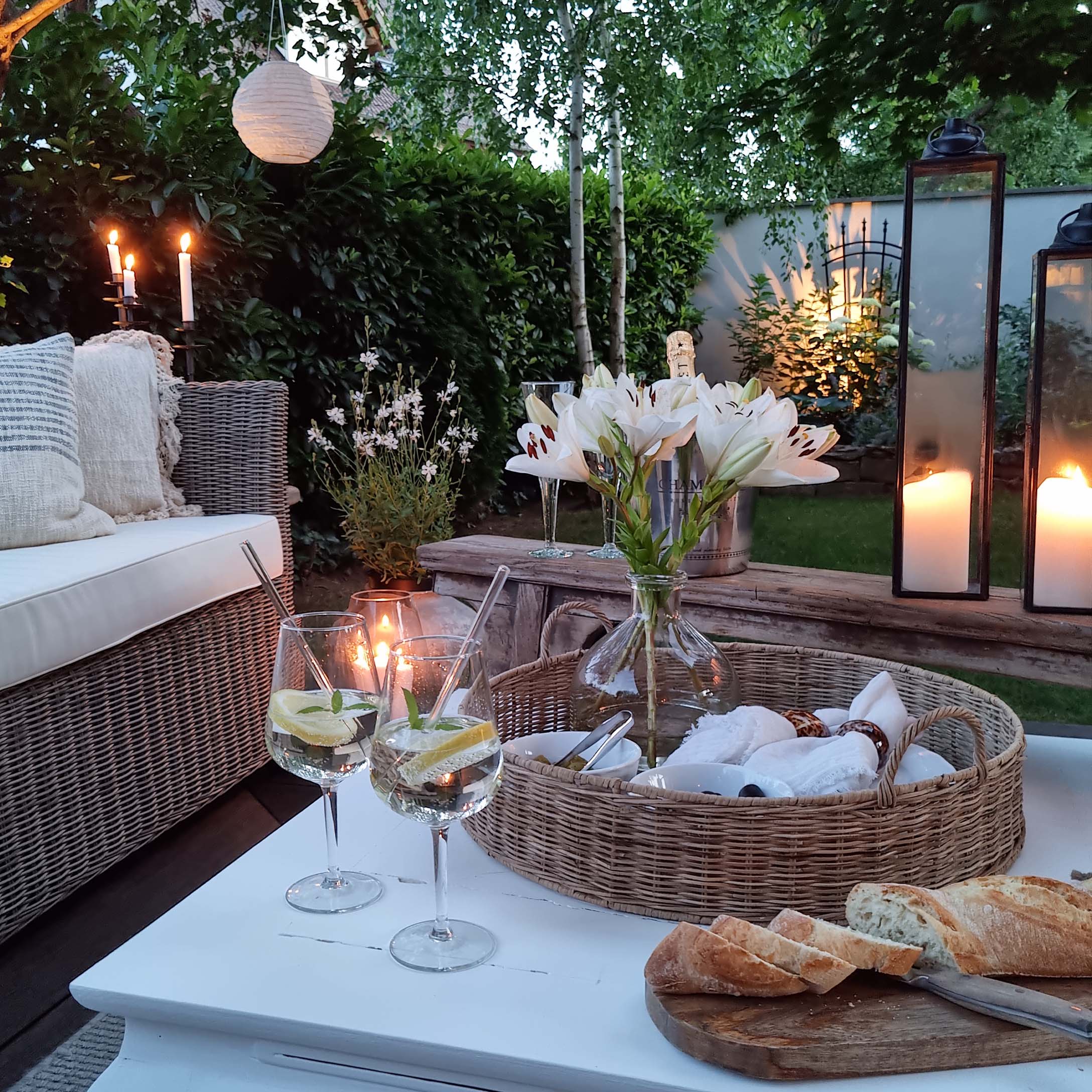 Jardin cosy avec canapé en osier, lys blancs et lanternes sur table en bois blanc