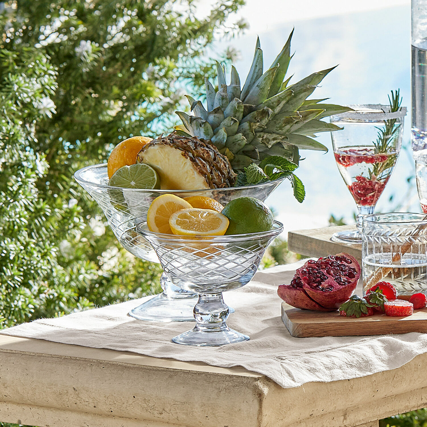 Lot de 2 coupes Kadence Coupes en verre sur pied avec fruits agrumes et ananas sur table claire en extérieur