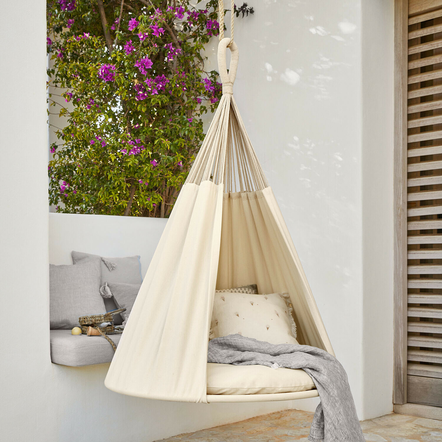 Hamac suspendu beige en coton avec coussins et couverture grise sur terrasse fleurie