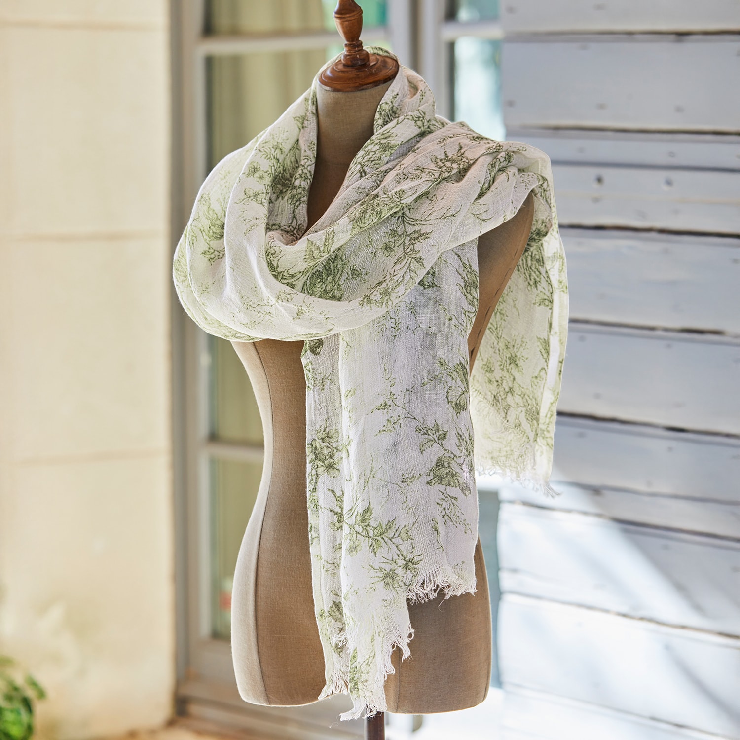 Écharpe légère à motif floral vert sur fond crème, présentée dans un jardin avec panier en osier