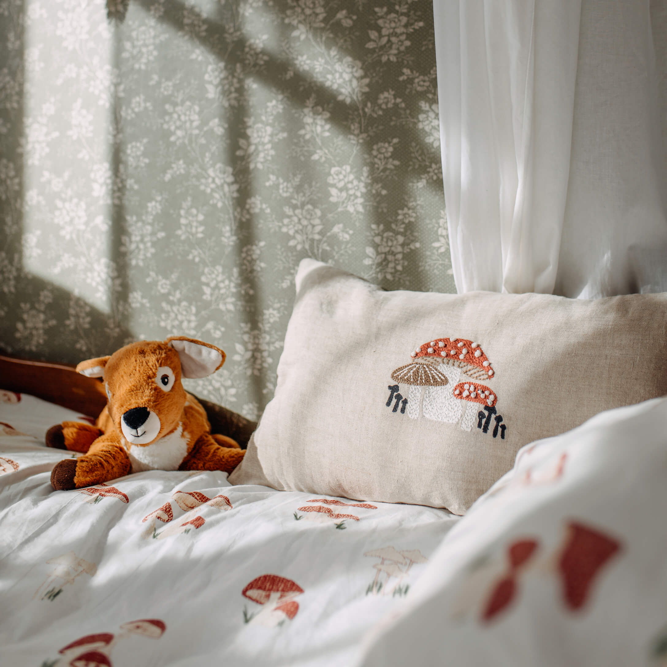 Coussin en lin beige brodé de champignons sur un lit avec une literie à motif champignons et une peluche cerf