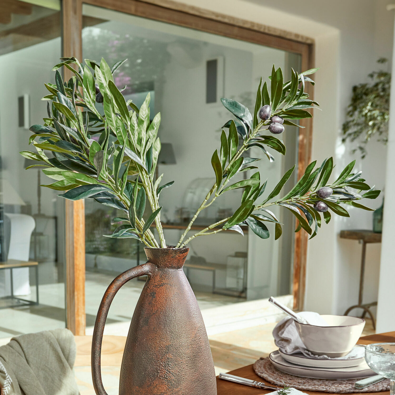 Lot de 2 branches d’olivier décoratives Olives Pichet rustique en métal couleur rouille avec branches d'olivier vertes sur une table à manger