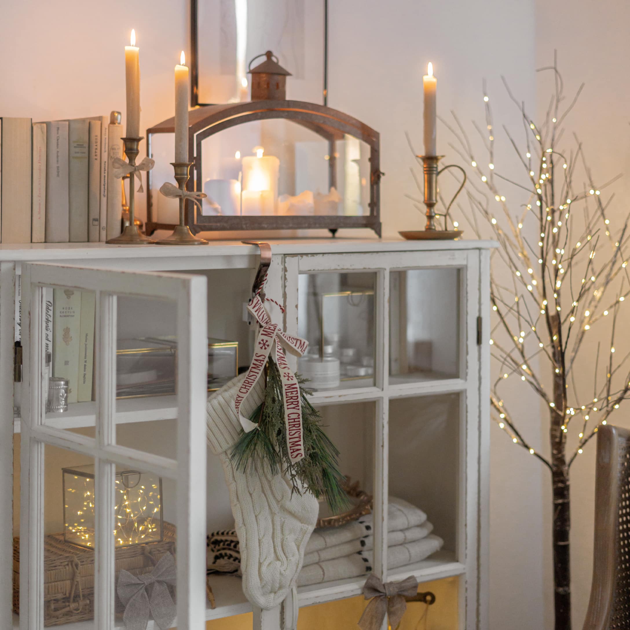 Chaussette de Noël blanche en tricot avec branches vertes sur une armoire blanche décorée de bougies et guirlandes lumineuses