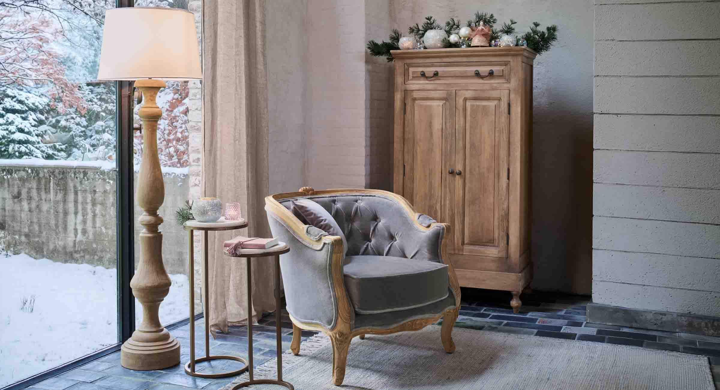 Coin cosy avec fauteuil en velours gris et lampe en bois près d'une fenêtre donnant sur un paysage enneigé