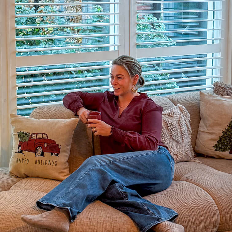 Femme en chemise bordeaux et jean se détend sur un canapé beige avec coussin décoratif rouge et message festif