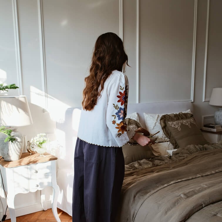 Femme en chemisier blanc à broderies colorées dans une chambre avec literie marron et lampe sur table de chevet