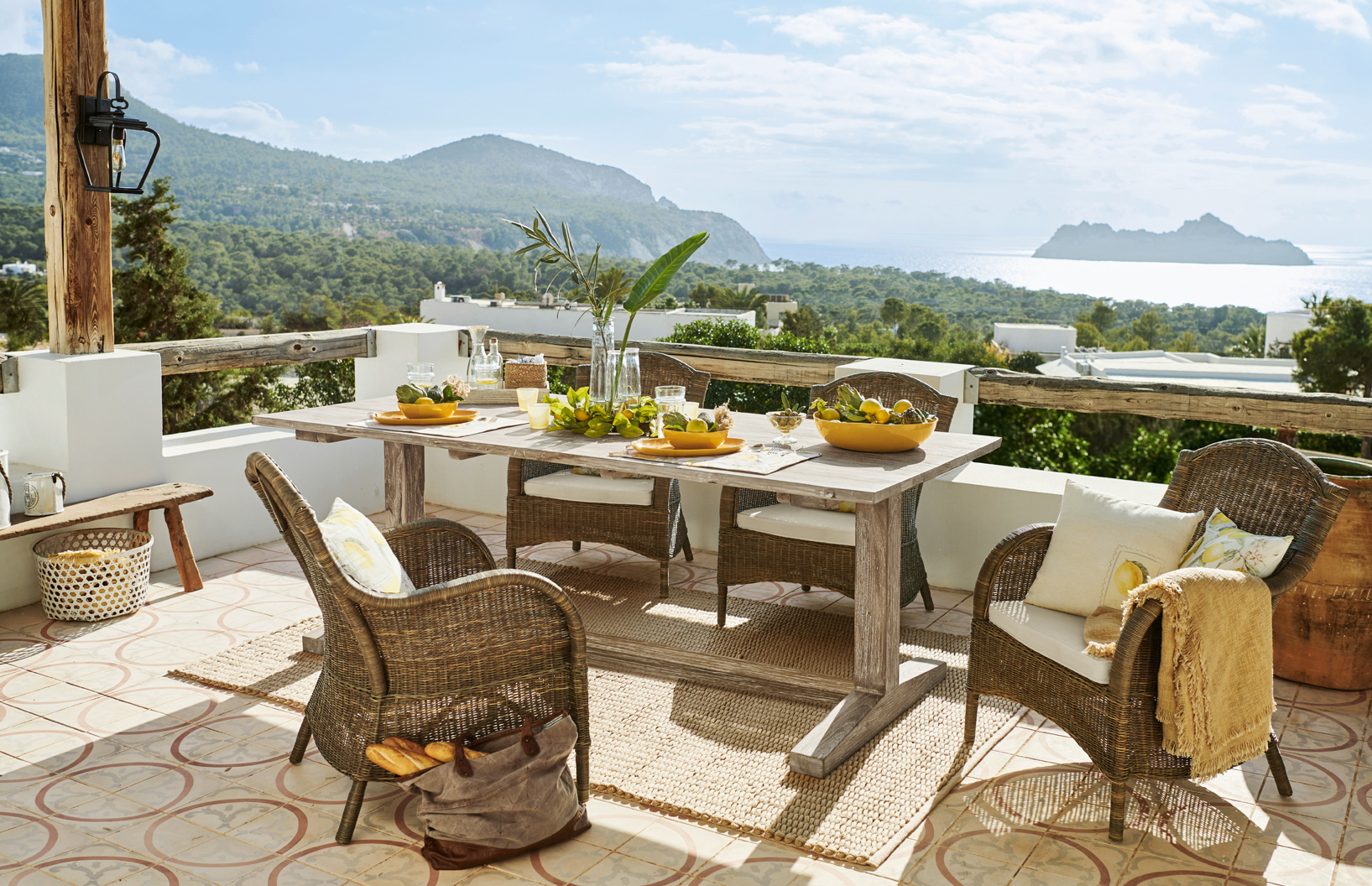 Terrasse avec mobilier en rotin et table en bois clair, vue sur mer et montagnes, style méditerranéen