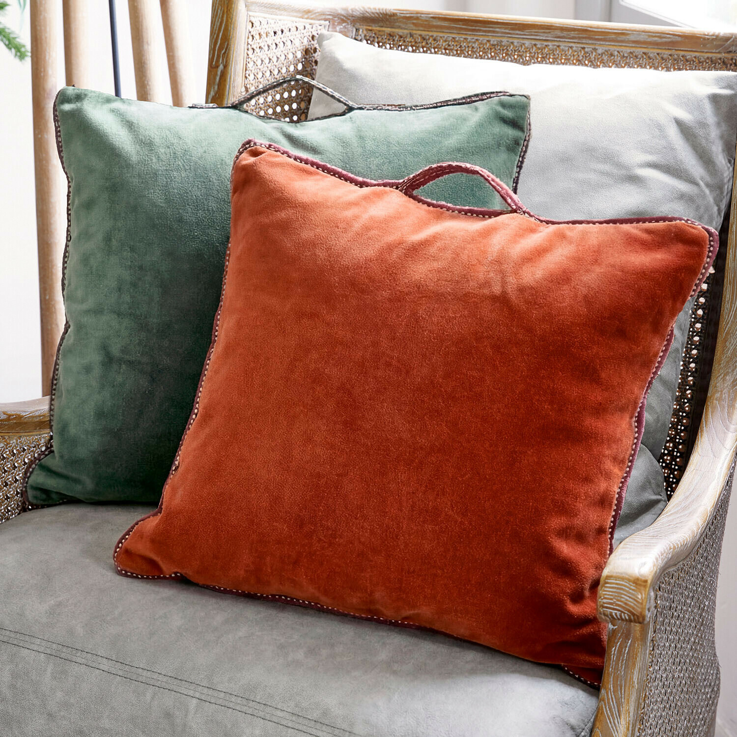 Lot de 2 coussins Olivienne Trois coussins décoratifs en velours vert, orange et gris sur un fauteuil en osier pour un confort élégant