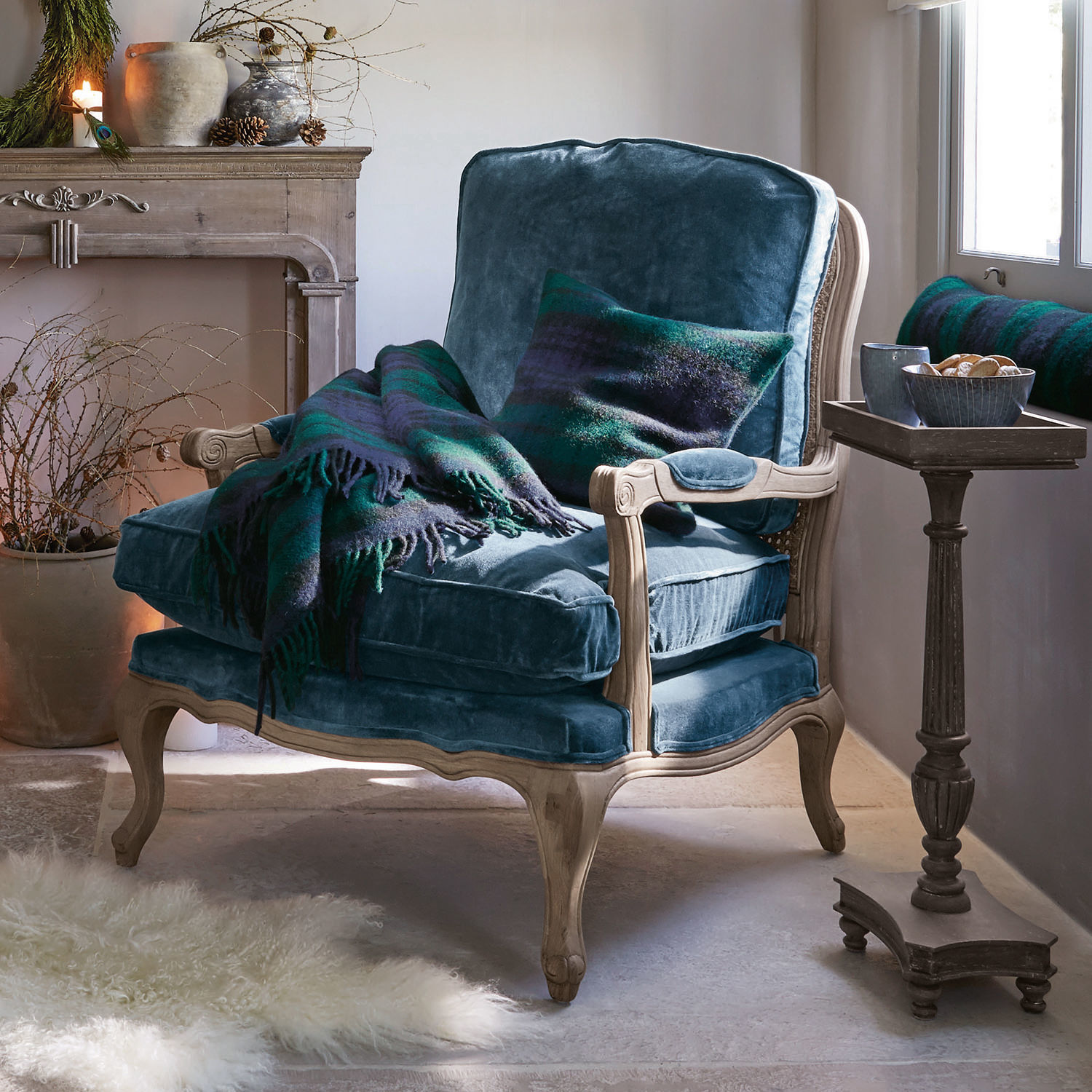 Fauteuil en velours bleu avec cadre en bois clair, plaid et coussin verts et bleus foncés