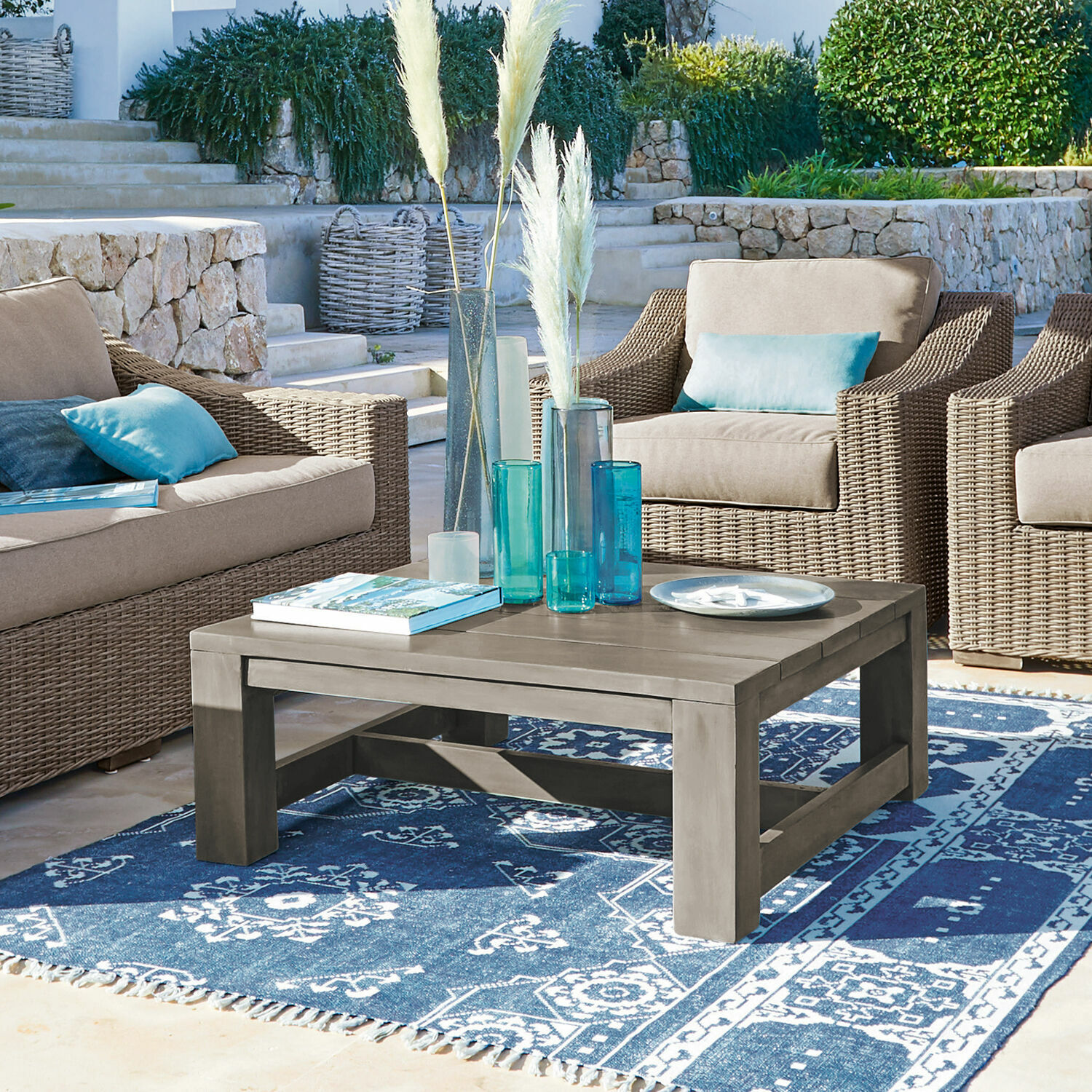 Table basse Malden Table basse en bois gris sur tapis bleu à motifs, entourée de fauteuils en osier avec coussins bleus.