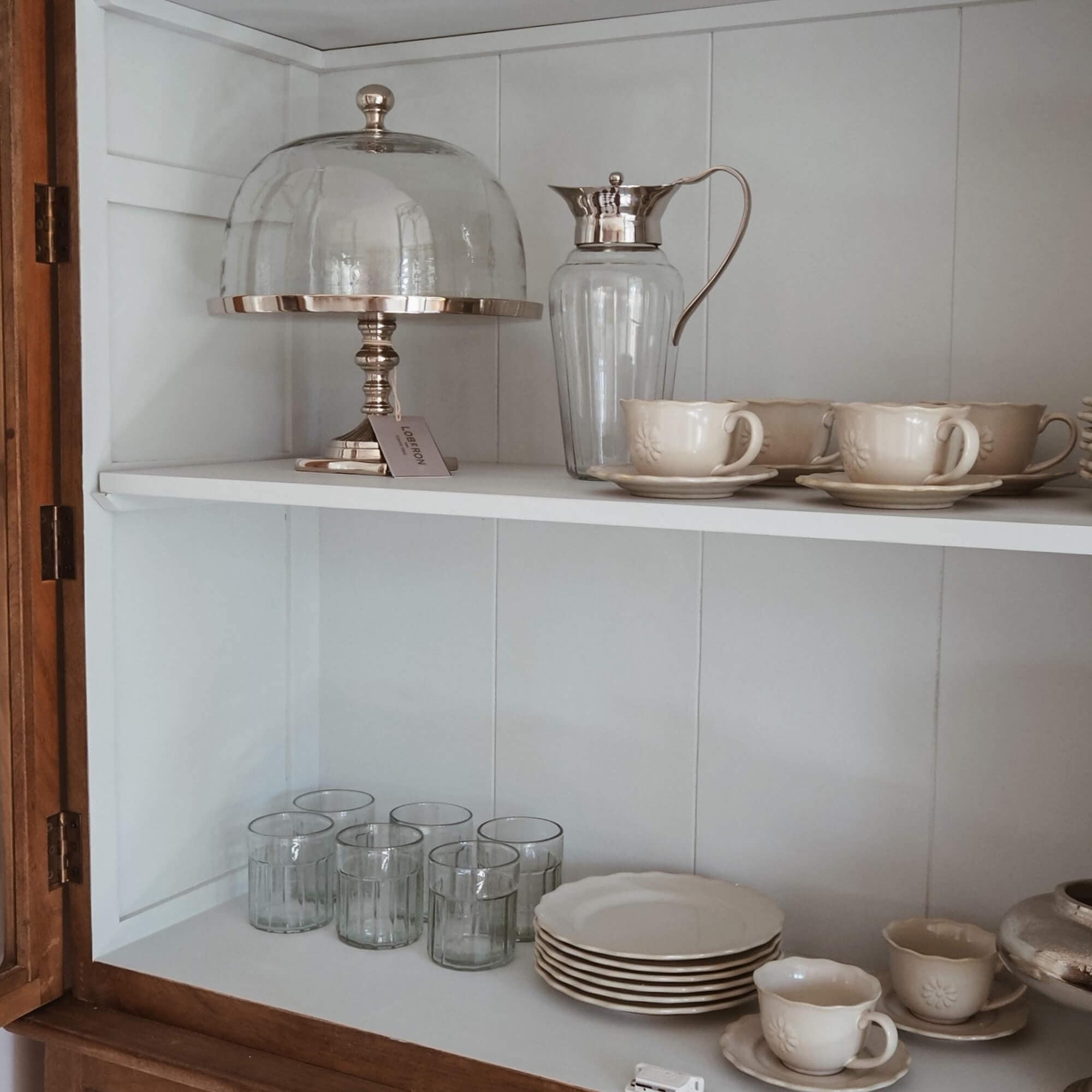 Ensemble de tasses et assiettes crème en céramique avec carafe en verre et cloche à gâteau dans une armoire blanche