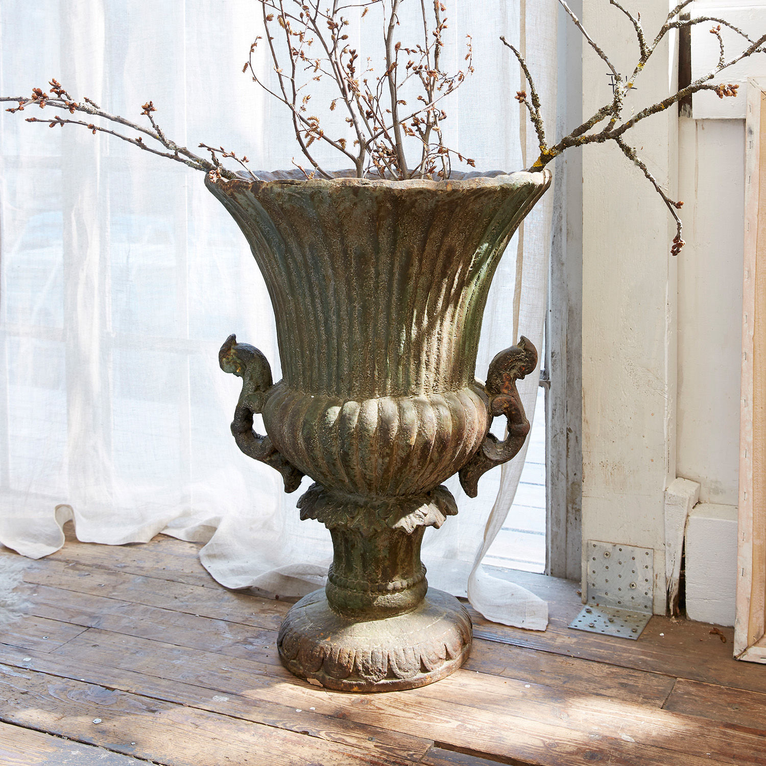 Amphore Estefanie Grand vase décoratif en métal vert patiné posé sur un sol en bois avec des branches sèches