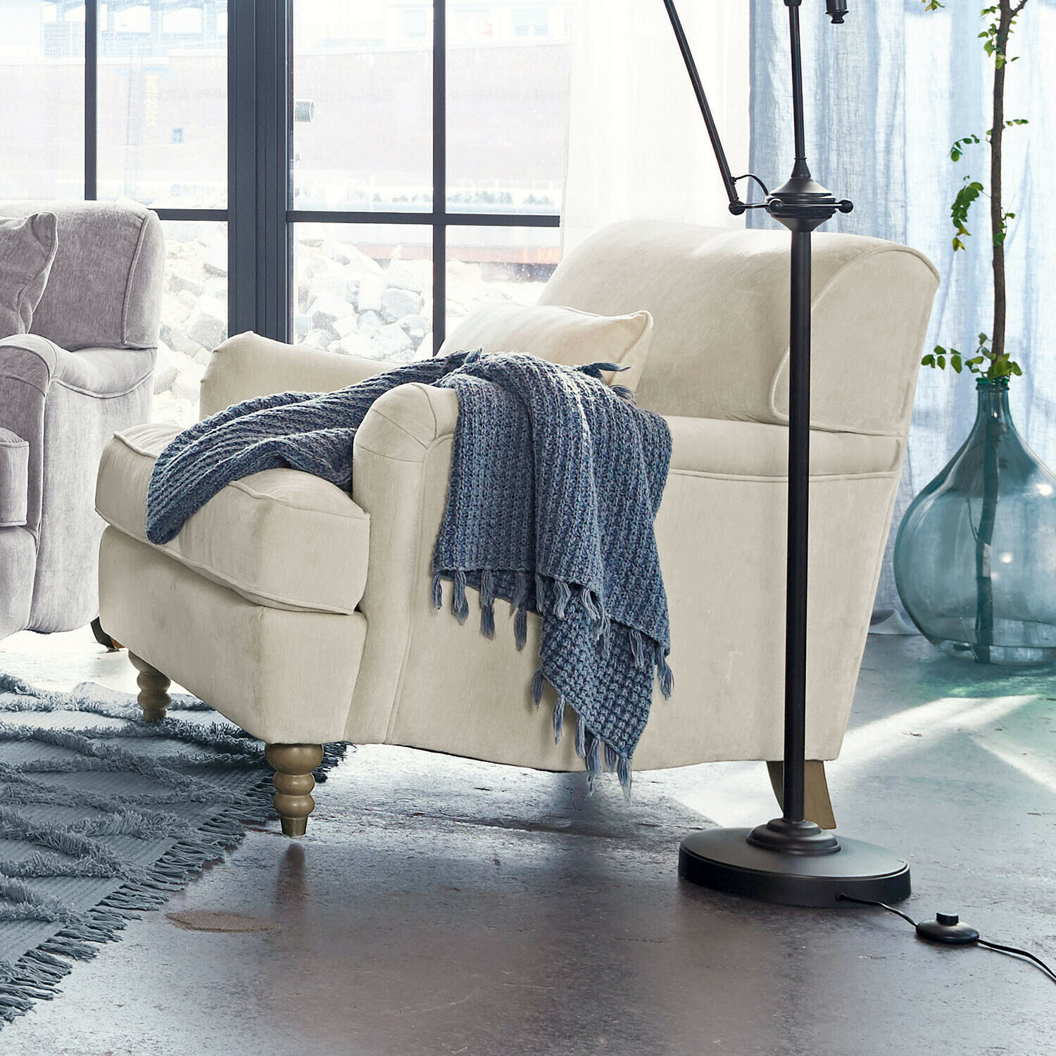 Fauteuil Isa Canapé beige en tissu avec pieds en bois et plaid bleu tricoté dans un salon lumineux moderne