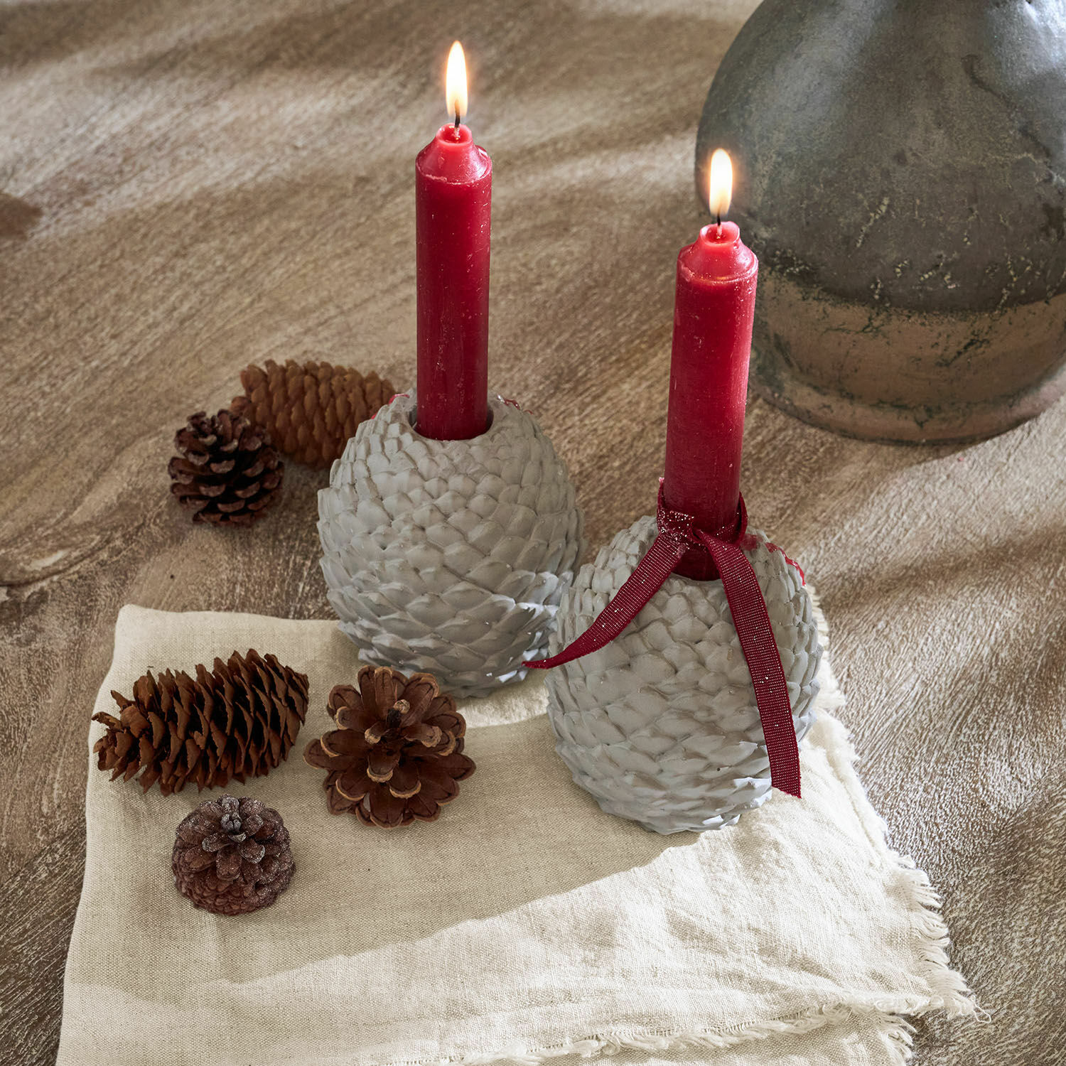 Lot de 2 bougeoirs Rianora Deux bougeoirs gris en forme de pomme de pin avec bougies rouges sur tissu en lin et pommes de pin