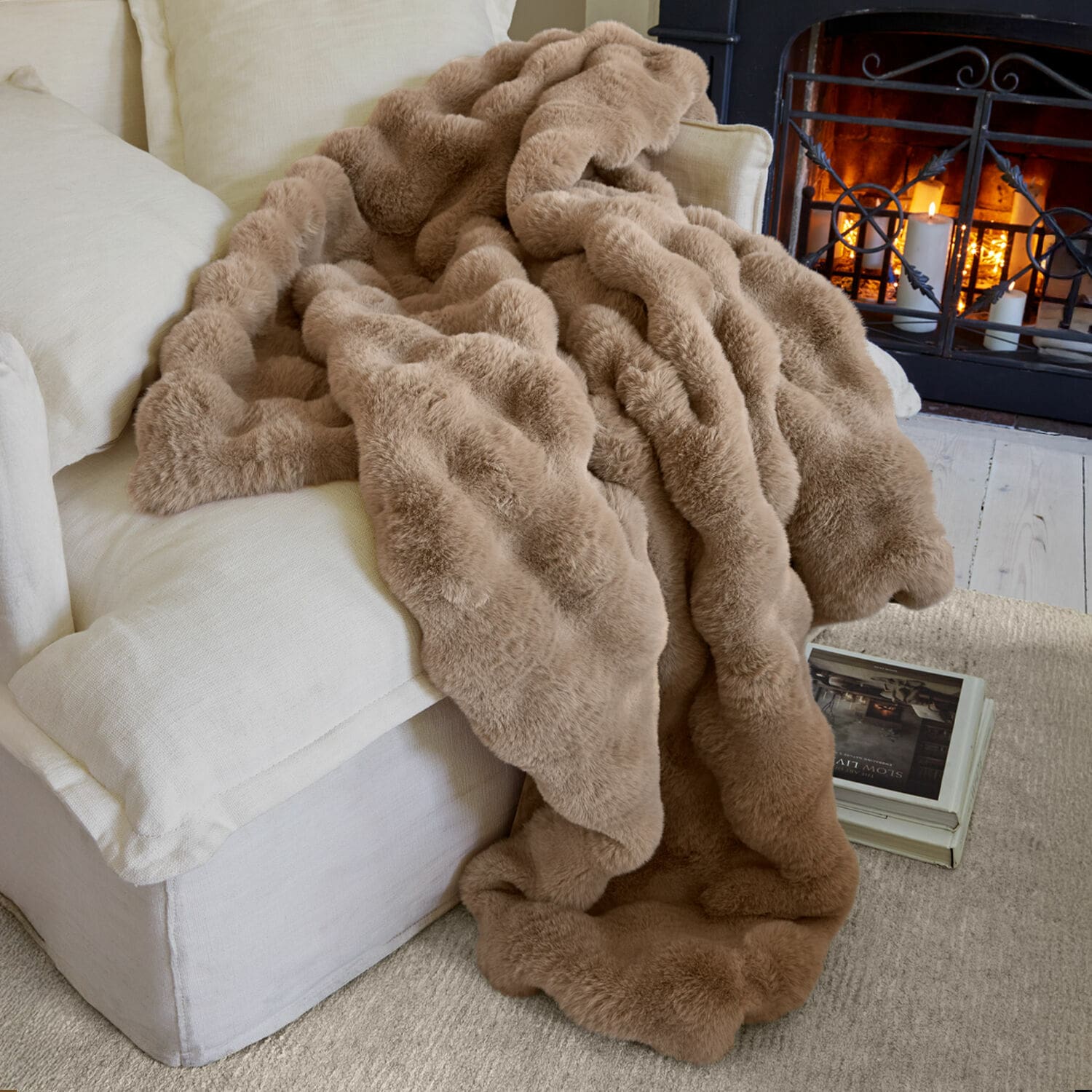 Plaid Zalina Plaid beige en fausse fourrure sur canapé clair près d'une cheminée allumée pour plus de confort