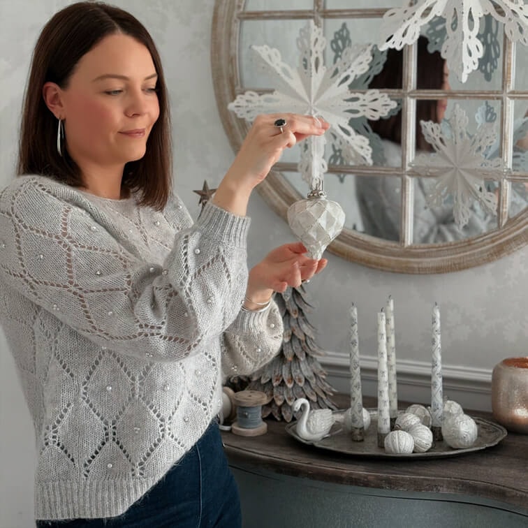 Femme en pull gris orné de perles tenant une décoration blanche de Noël dans un intérieur chaleureux avec décorations hivernales