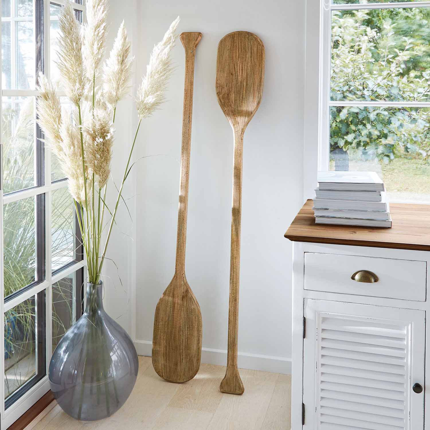 Deux pagaies décoratives en bois naturel appuyées contre un mur blanc près d'une commode et d'un vase avec des herbes de la pampa