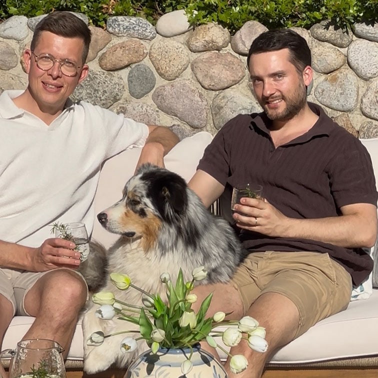 Deux hommes assis sur un canapé extérieur avec un chien berger australien et un bouquet de tulipes blanches.