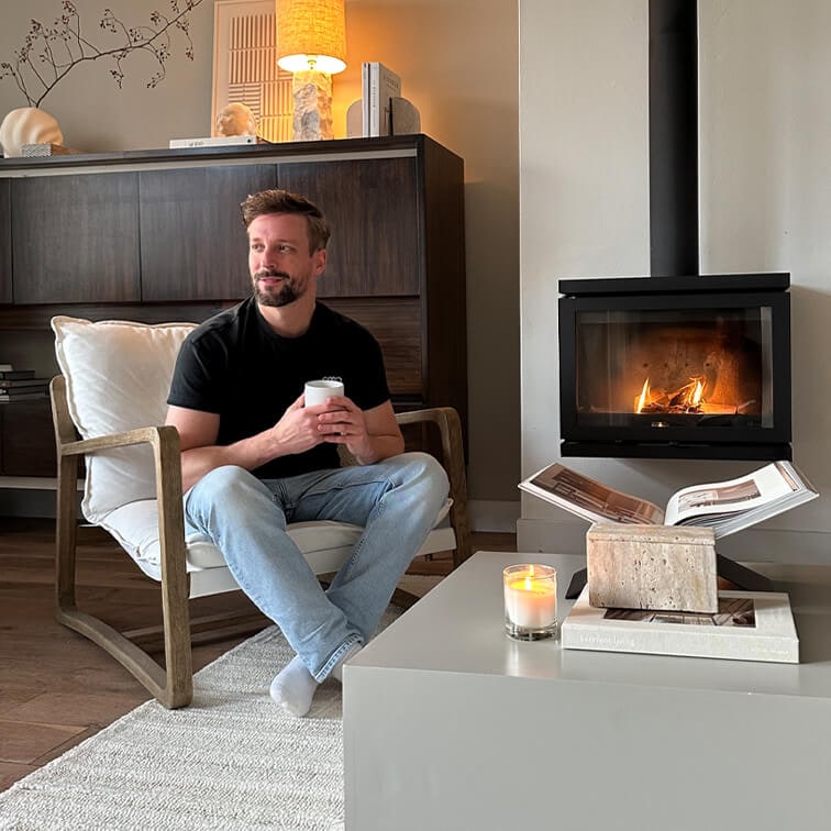 Homme assis sur un fauteuil en bois avec coussin blanc, tenant une tasse, cheminée et bougie sur la table