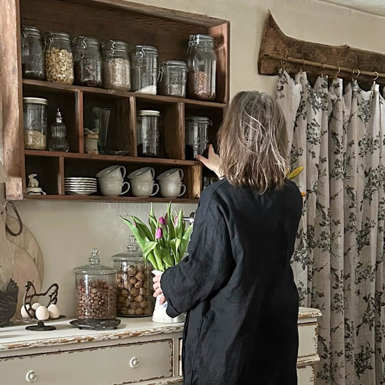 Femme en robe noire attrapant un bocal sur une étagère en bois dans une cuisine rustique avec tulipes