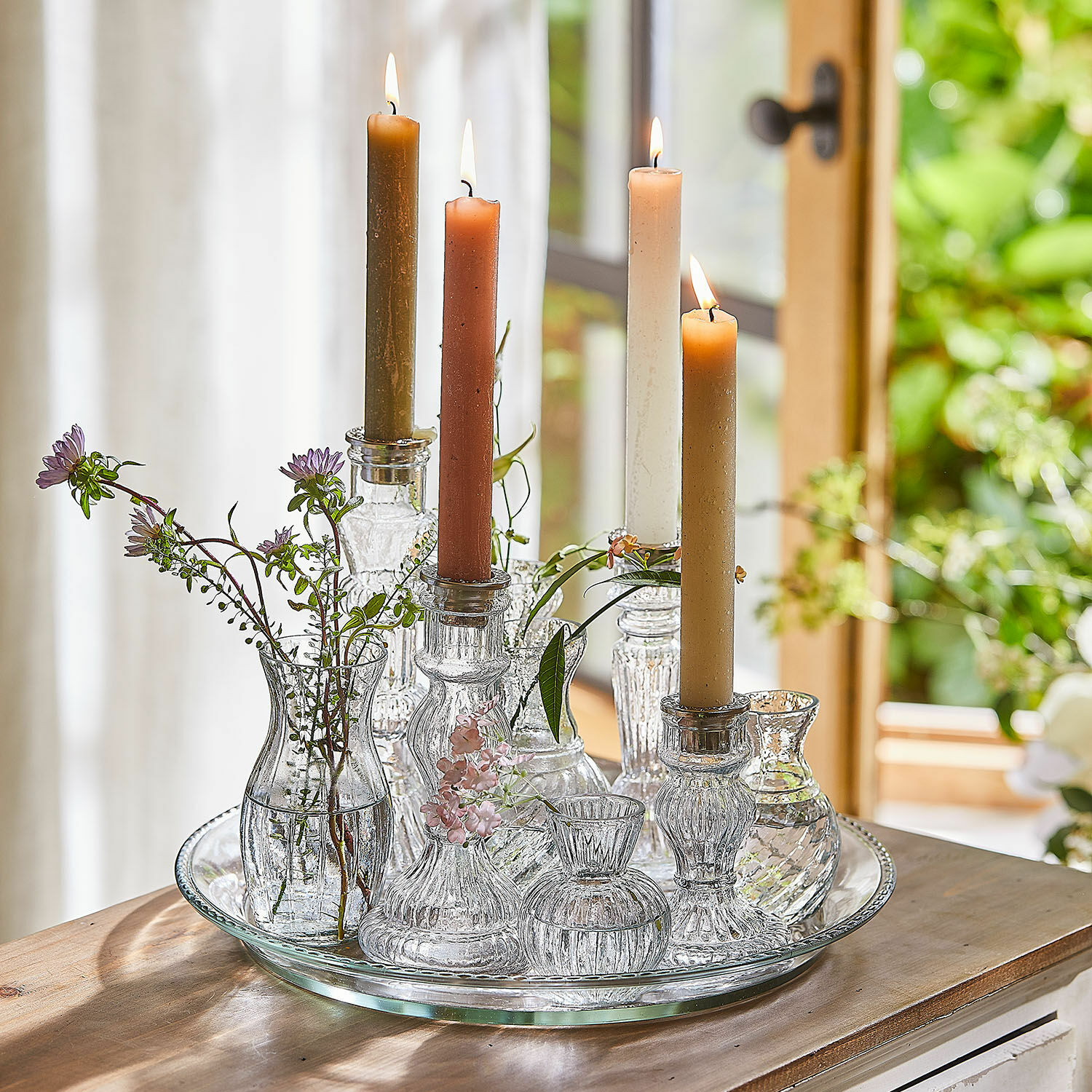 Plateau avec bougeoirs et vases Hilevor Bougeoirs et petits vases en verre avec bougies colorées et fleurs sur un plateau en verre sur une table en bois