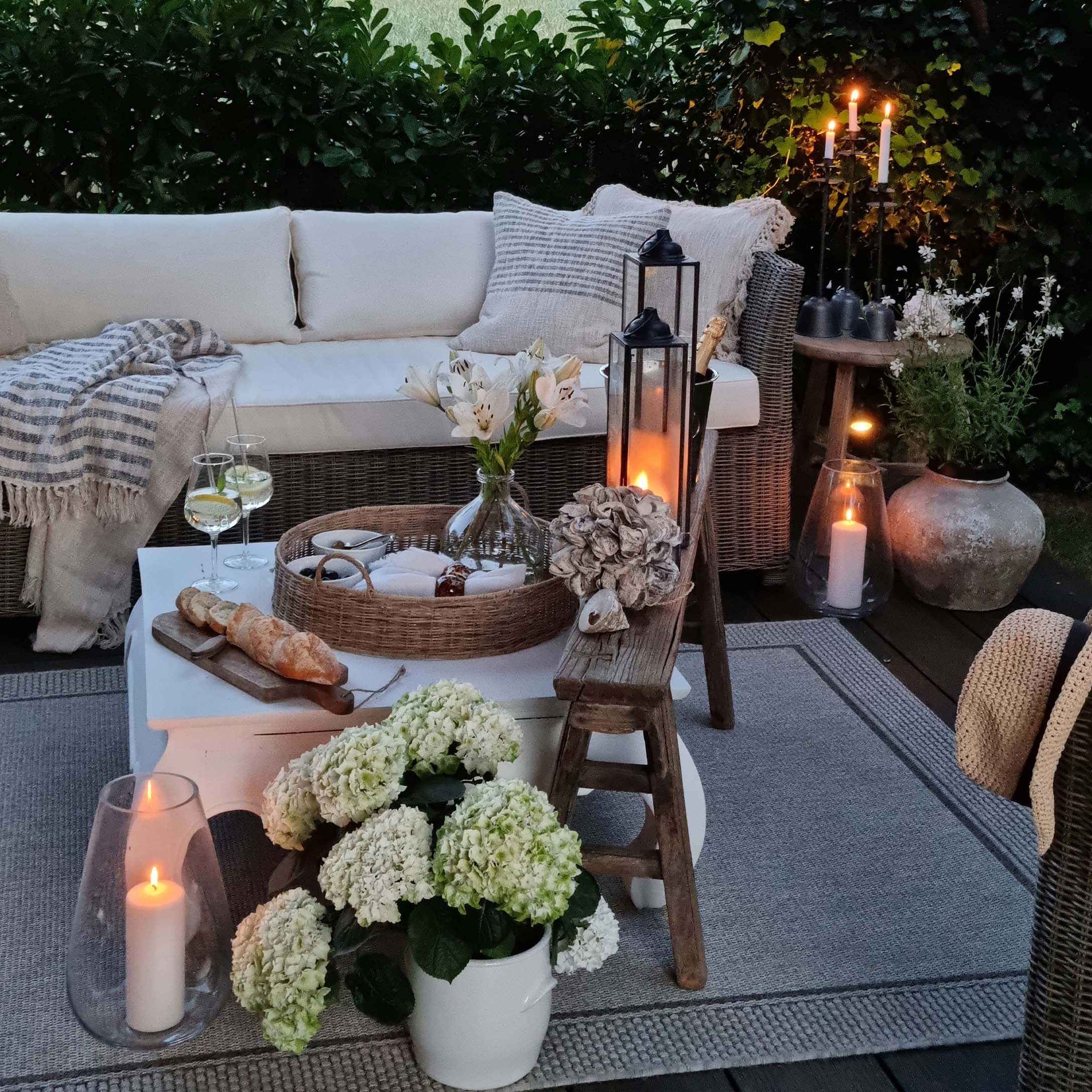 Coin jardin chaleureux avec canapé en osier, coussins blancs et bougies sur terrasse en bois