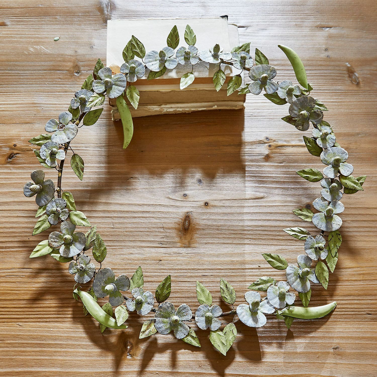 Couronne Cecilia Couronne décorative en métal avec feuilles vertes et fleurs blanches posée sur une table en bois